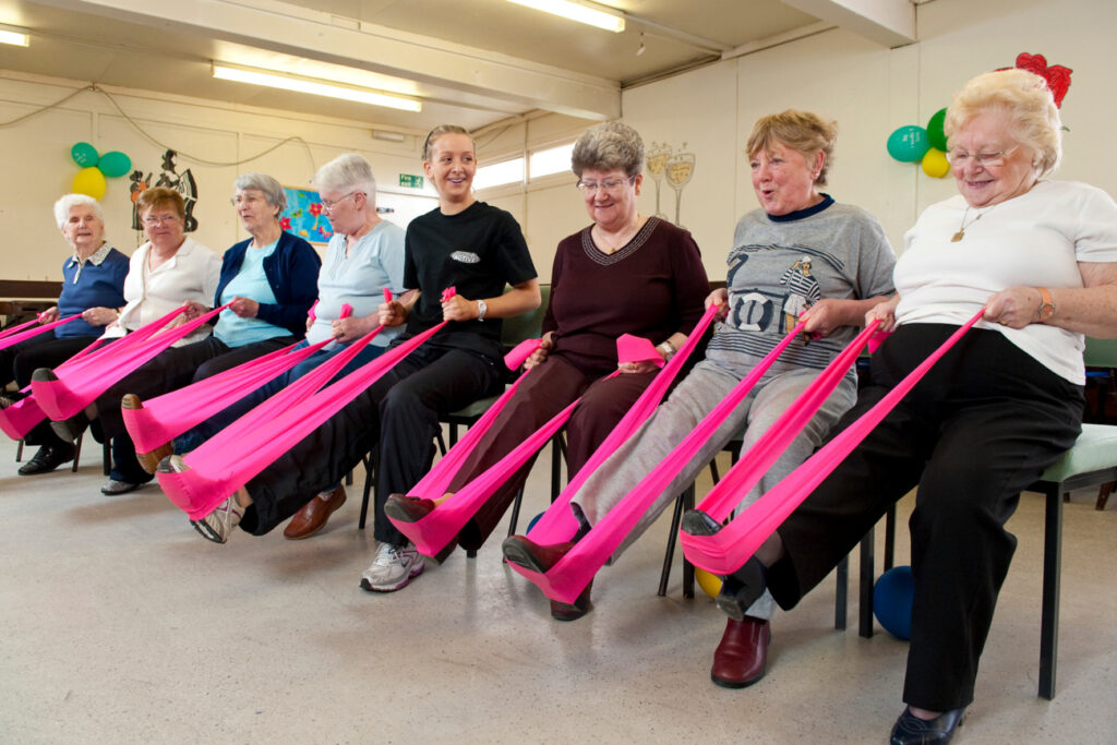 Seven women are sitting in a row on chairs. They are each holding a bright pink resistance band which is looped around their right foot, and they are using it to stretch