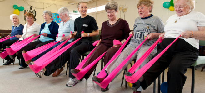 Seven women are sitting in a row on chairs. They are each holding a bright pink resistance band which is looped around their right foot, and they are using it to stretch